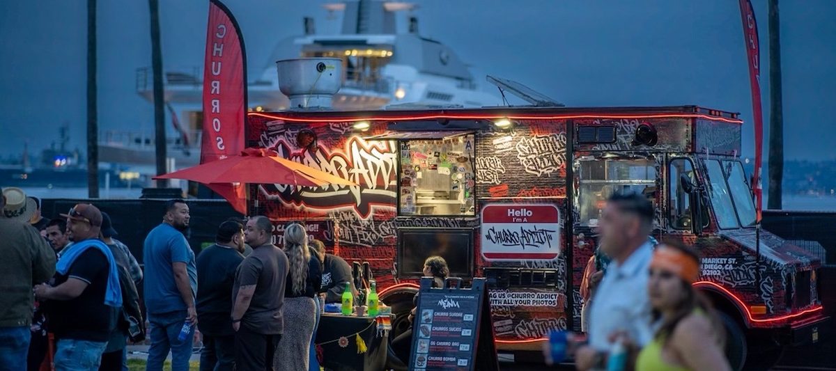 People lined up to order from red and black food truck with graffiti logo in park with yachts and sea in background. Churro Addict Food Truck, Best Food Trucks San Diego.
