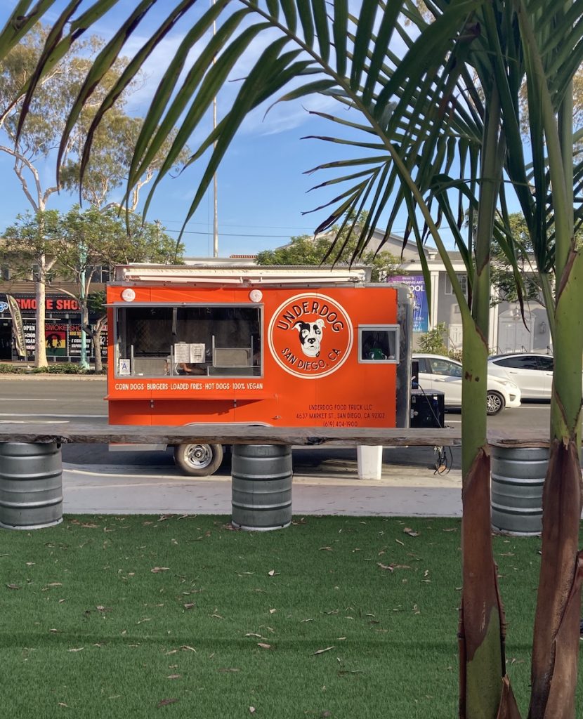 Small red food truck outside surrounded by palm tree on sunny day. Underdog Food Truck.