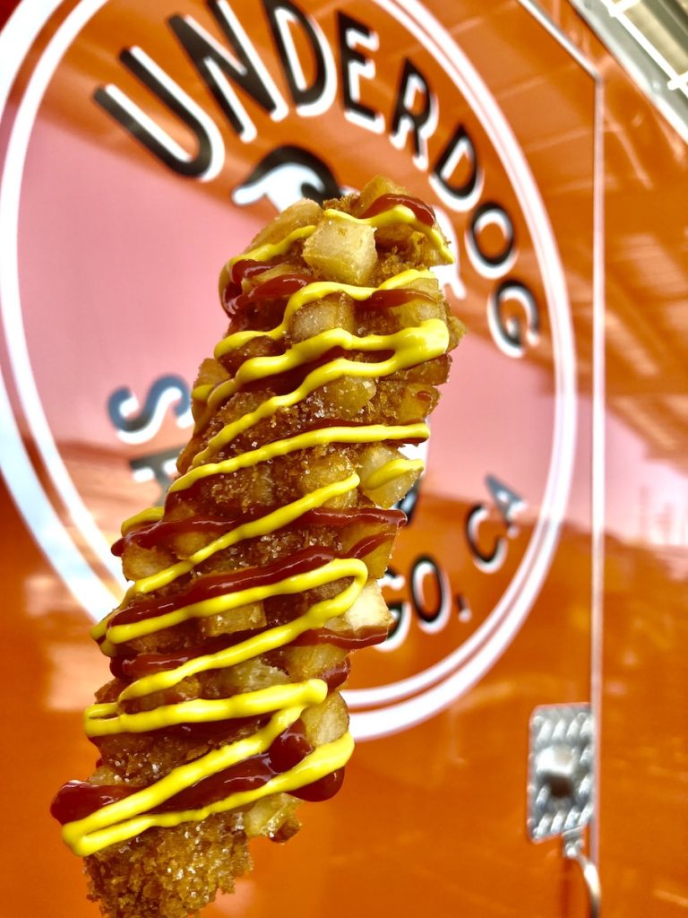 Close up of corn dog covered in ketchup and mustard with red food truck in background. Underdog Food Truck, Food Trucks San Diego.